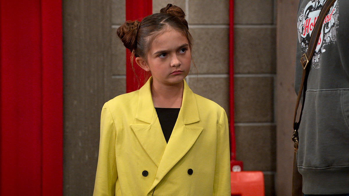 &Quot;Young Girl With Double Bun Hairstyle, Wearing A Bright Yellow Blazer, Stands Indoors Next To An Adult In Casual Clothing, With A Red Wall And Pipes In The Background, Looking Slightly Confused Or Skeptical.&Quot;