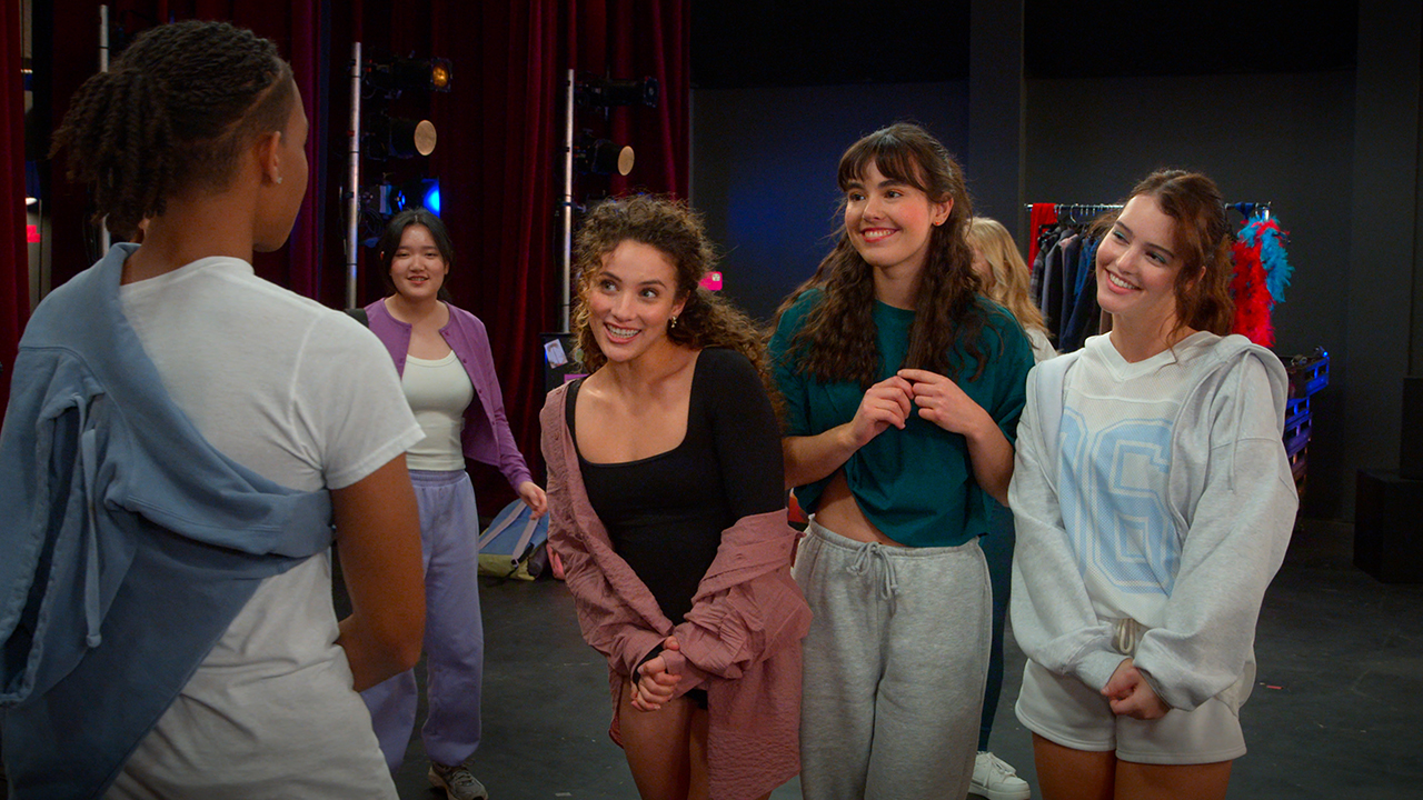 &Quot;A Group Of Young Adults In Casual Dancewear Stand Together Smiling And Talking On A Theater Stage, With Backstage Props And Red Curtains Visible In The Background.&Quot;