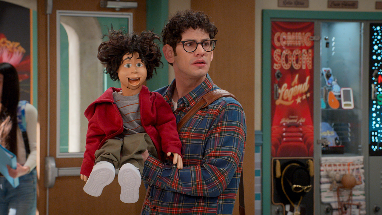 &Quot;A Man With Curly Hair And Glasses Stands In A School Hallway Holding A Puppet Wearing A Red Jacket. There Are Lockers, Posters, And A Vending Machine In The Background, Suggesting A Casual And Lively Academic Environment.&Quot;