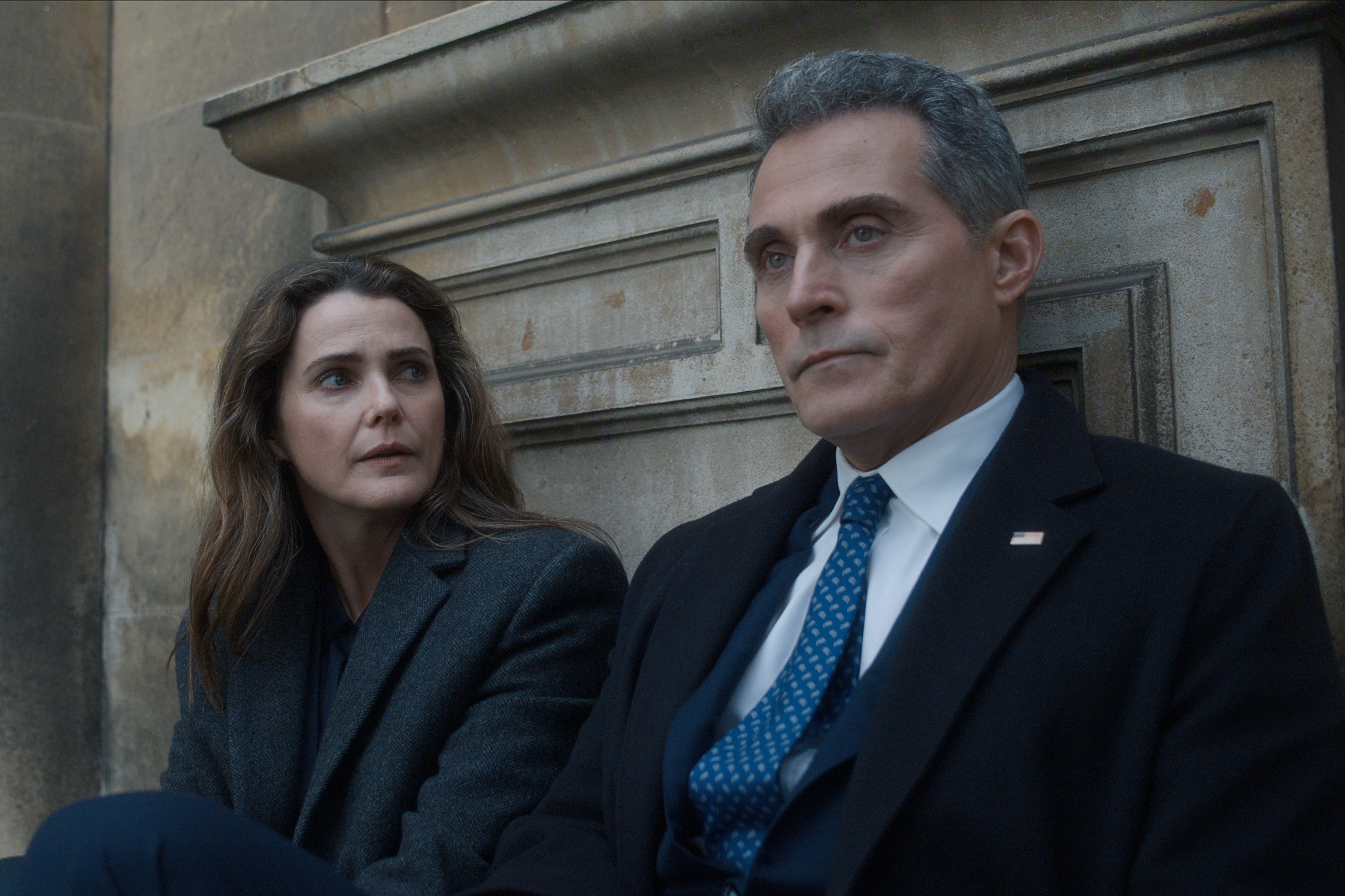 "Two people wearing formal coats are sitting on the outdoor stone steps, looking serious and deep in conversation, with an ancient stone wall in the background. The man wears a suit and tie with an American flag pin, indicating a political or formal context."