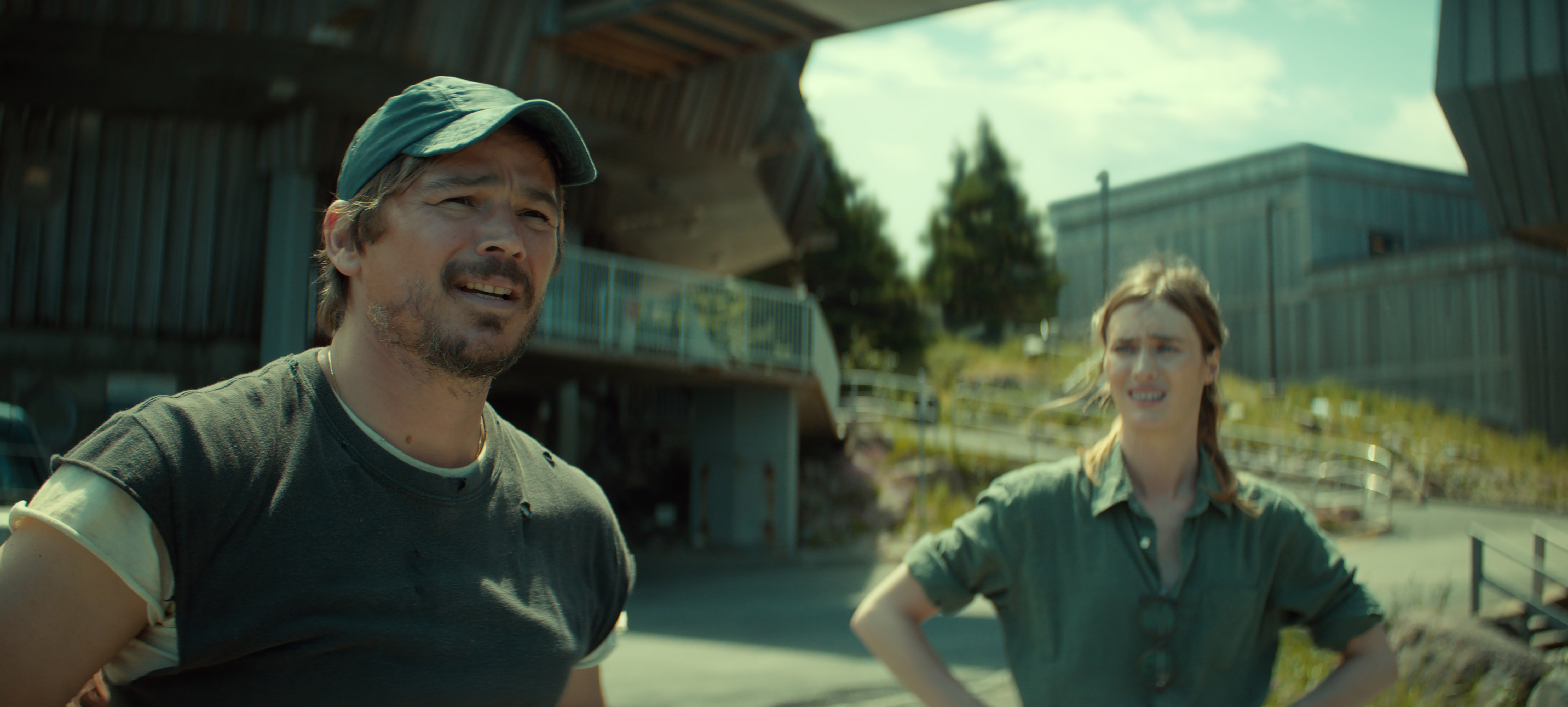 "Two people stand outdoors near modern industrial buildings on a sunny day. The man wears a cap and casual clothes, while the woman stands with her hands on her hips, both looking in the same direction."