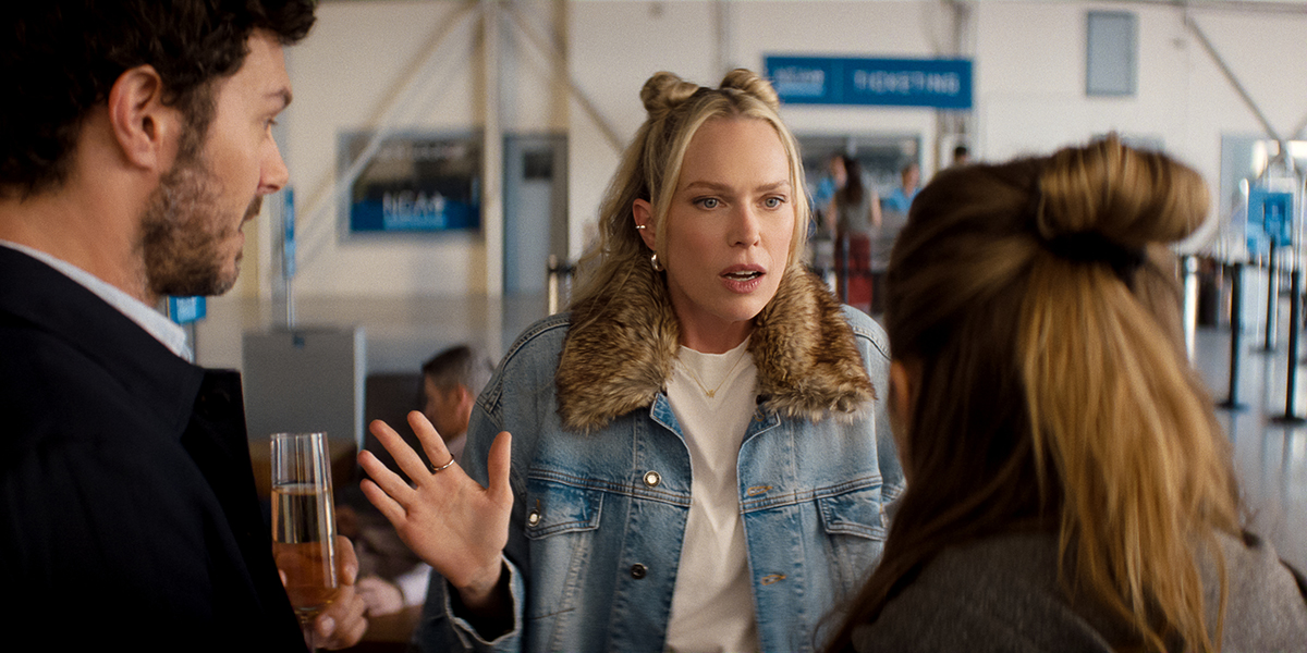 &Quot;Three People Talking In An Airport Terminal, One Woman In A Jean Jacket With Fur Collar Gesturing Animatedly, Appearing To Explain Something To A Man Holding A Drink And A Woman With Her Back To The Camera. Signs And Check-In Lines In Background.&Quot;