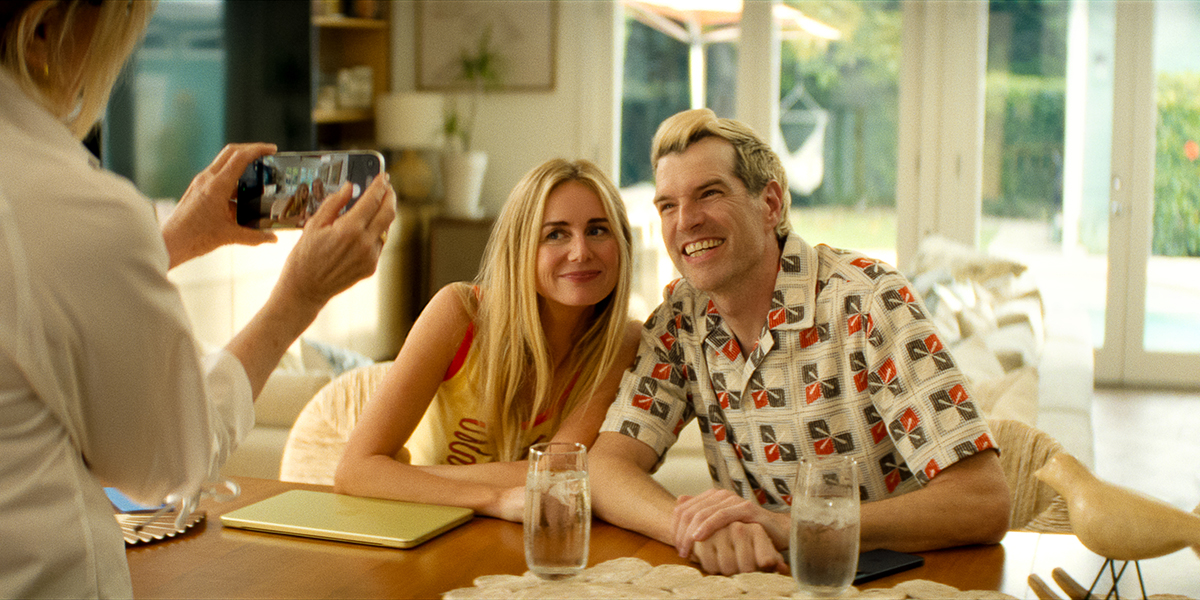 &Quot;A Smiling Couple Sits At A Table Indoors, Holding Hands And Posing For A Photo Taken By Another Person. Bright, Cozy Setting With Large Windows, Plants Outside, Two Glasses Of Water, And A Yellow Tablet On The Table.&Quot;
