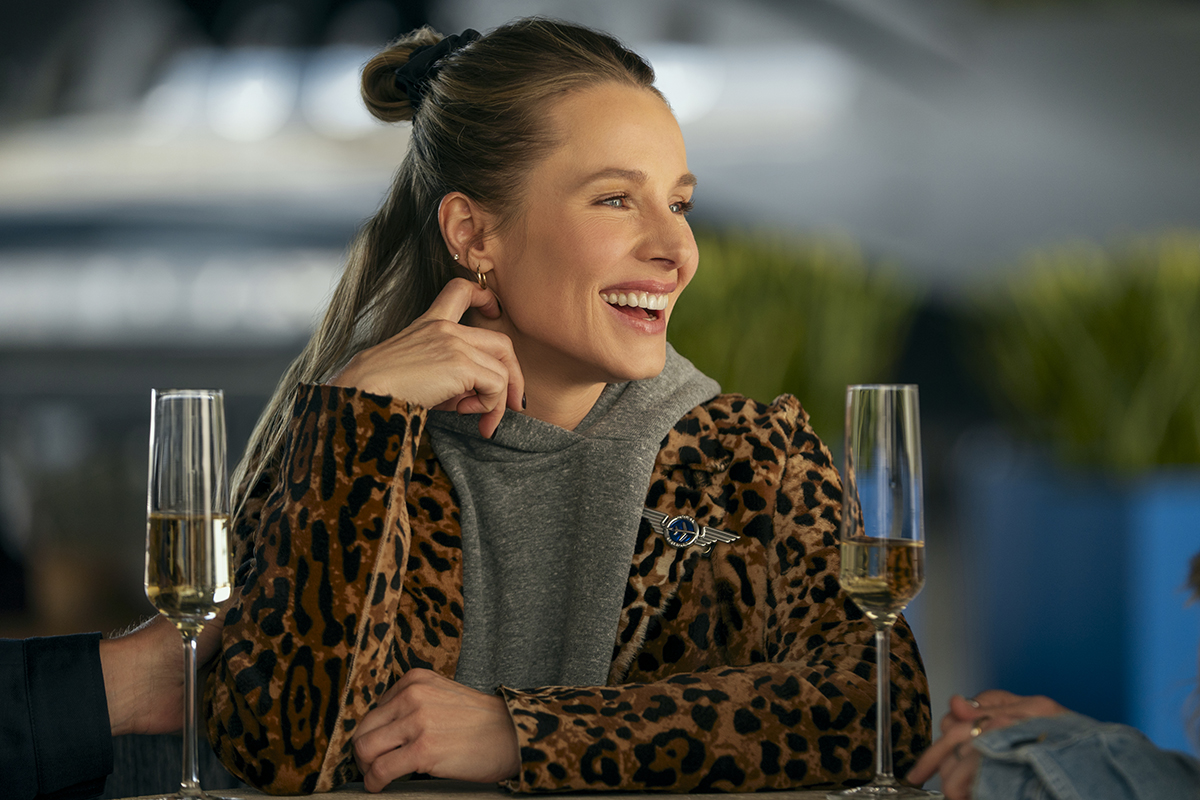 &Quot;Woman In Leopard Print Jacket Sitting At A Table, Smiling And Talking With Others, Two Glasses Of Champagne In Front Of Her, Modern Indoor Setting With Blurred Background And Plants.&Quot;