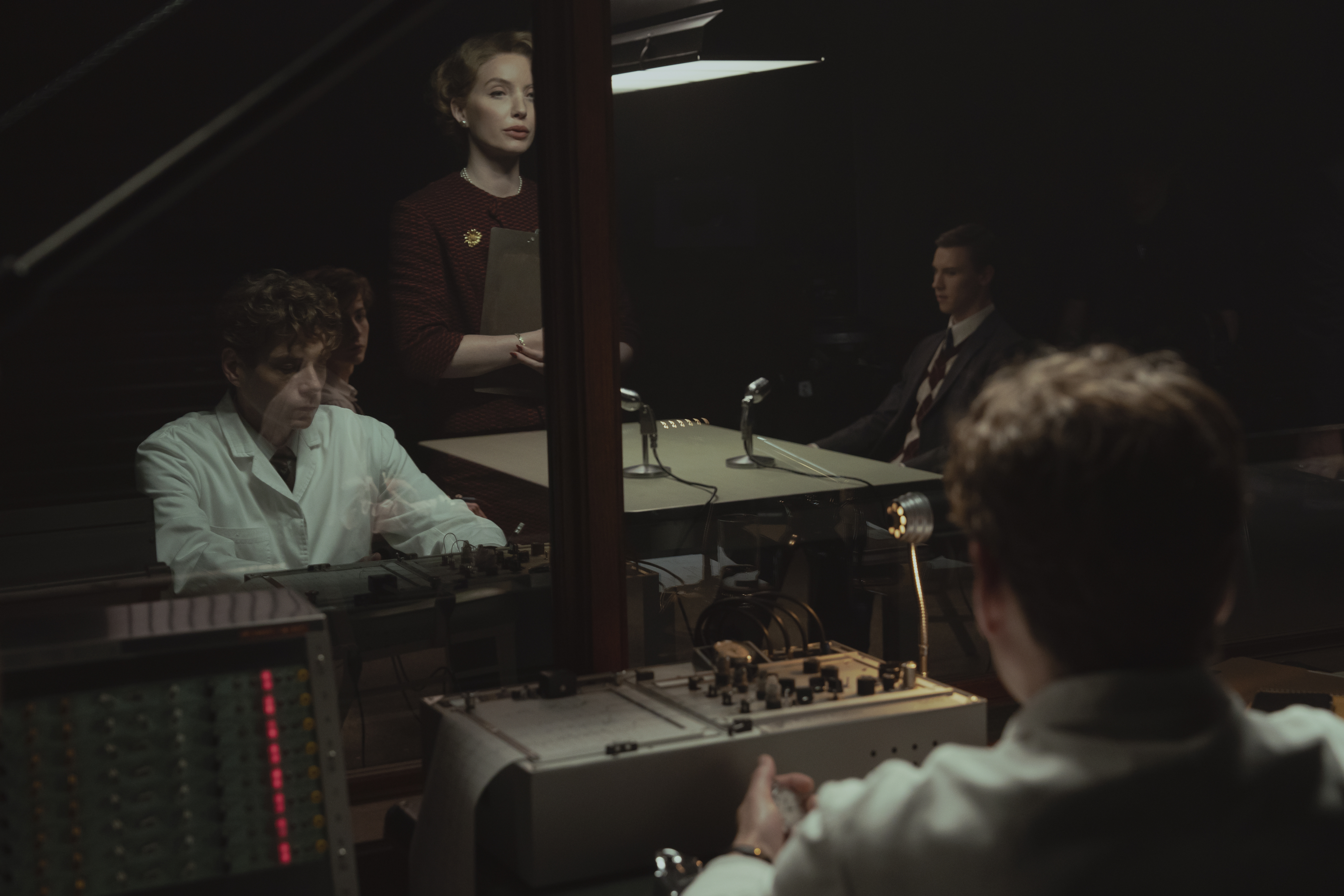 &Quot;A Group Of People In A Dimly Lit Room, Some In Lab Coats, Appear To Be Conducting Or Observing An Experiment Or Interrogation Through Glass, With Vintage Recording Equipment And Microphones On The Desk.&Quot;