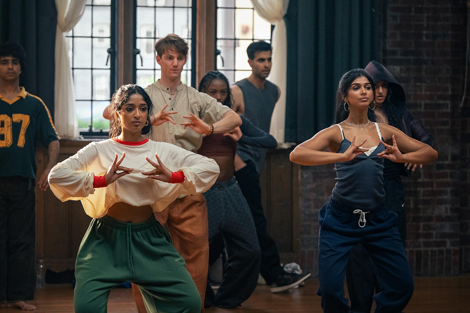 &Quot;A Diverse Group Of Young Adults Practicing Dance In A Studio With Wooden Floors, Brick Walls, And Large Windows. The Two Women In The Front Are Focused On Their Hand Movements, Suggesting A Choreographed Routine.&Quot;