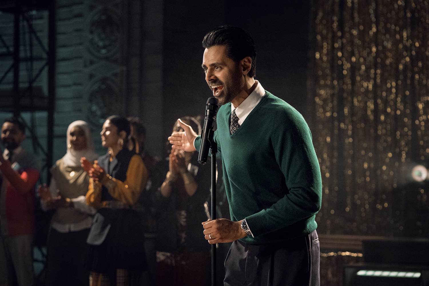 &Quot;Man In Green Sweater Speaks Into Microphone On Stage, With An Audience Of People Clapping And Watching In A Warmly Lit, Festive Indoor Environment With Golden Decorations In The Background.&Quot;