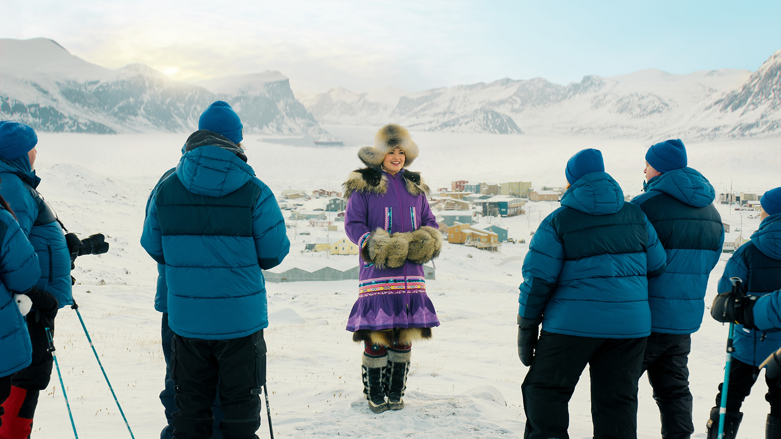 &Quot;A Woman In Traditional Fur-Trimmed Clothing Stands Smiling In Snowy Arctic Landscape, Surrounded By Five People In Blue Winter Jackets, With A Mountainous Village In The Background Under A Bright Sky.&Quot;