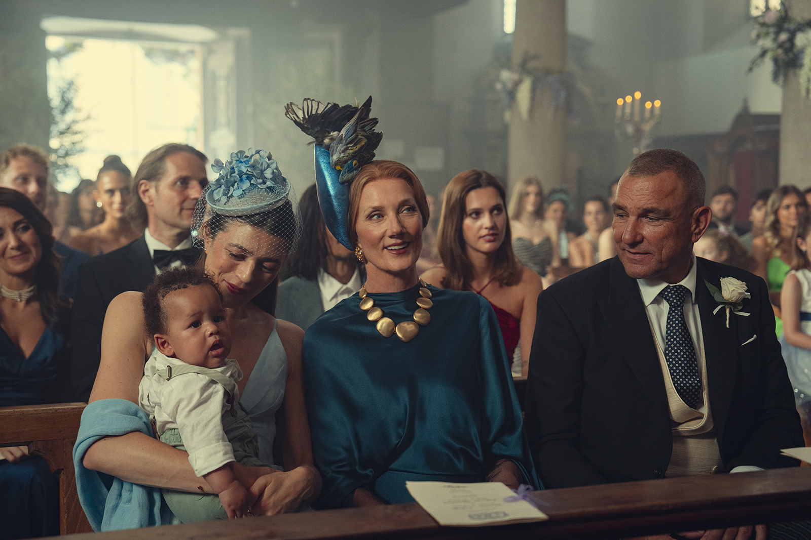 &Quot;People Dressed Formally Sitting In Pews At A Wedding Ceremony Inside A Church, With Women Wearing Hats And A Woman Holding A Baby; Candles And Floral Decorations Are Visible In The Background.&Quot;