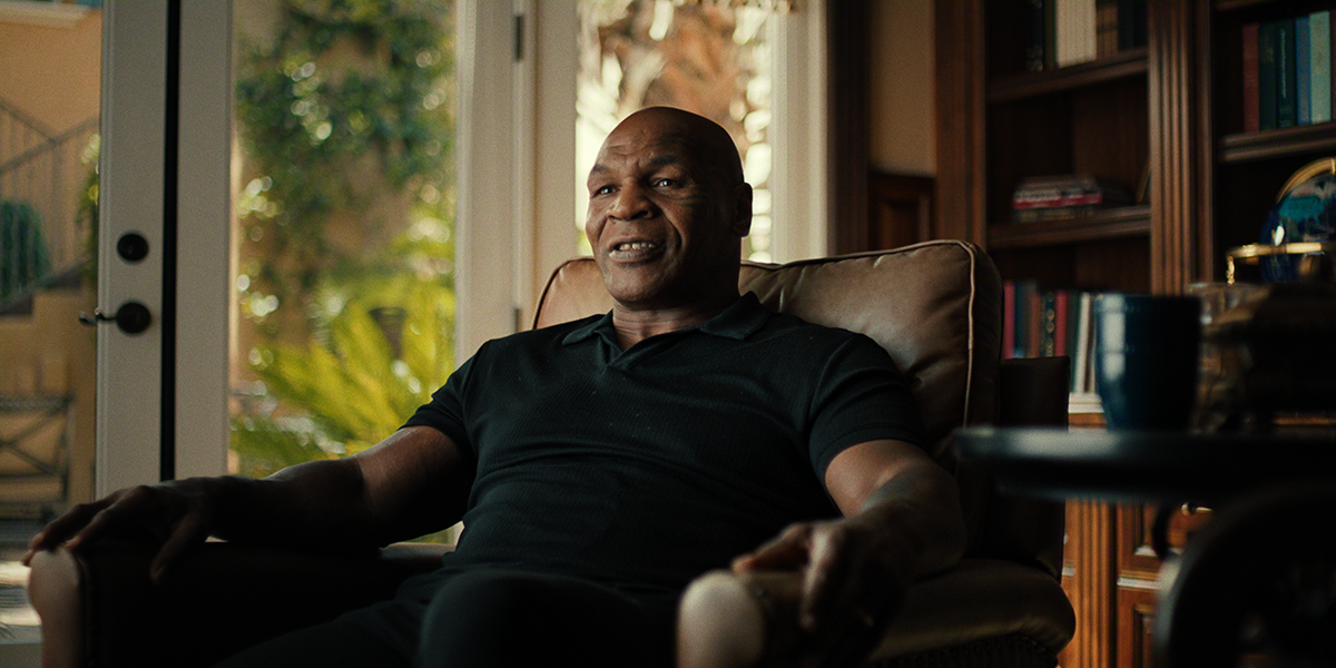 &Quot;A Man Sitting In A Cozy Armchair In A Warmly Lit Home, Surrounded By Bookshelves And Large Windows With Sunlight And Plants Visible Outside, A Coffee Table With A Mug In The Foreground.&Quot;