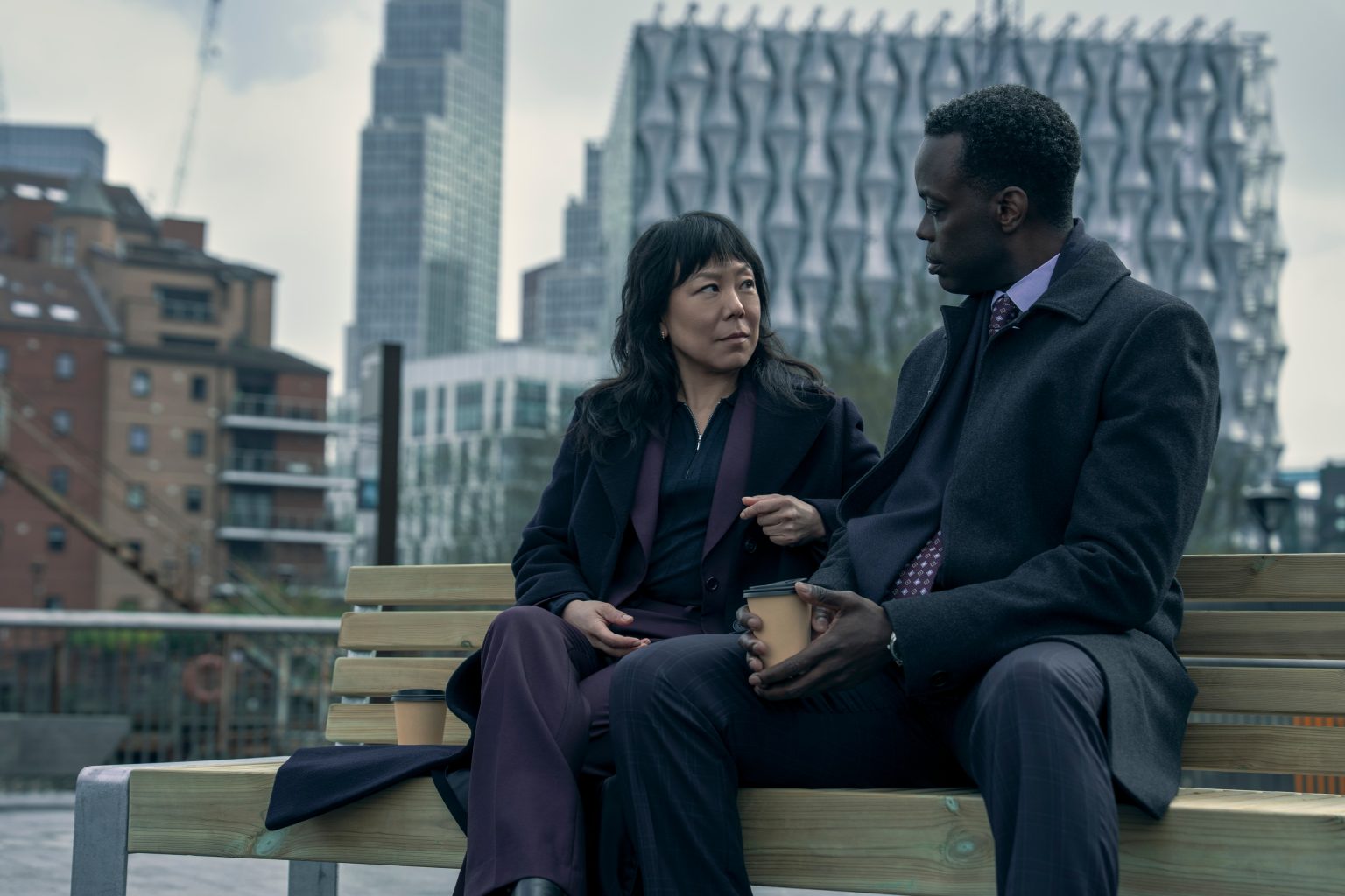 &Quot;Two People In Formal Attire Sit On A Bench Outdoors In A City, Modern Buildings With Unique Architecture In The Background, Having A Serious Conversation.&Quot;