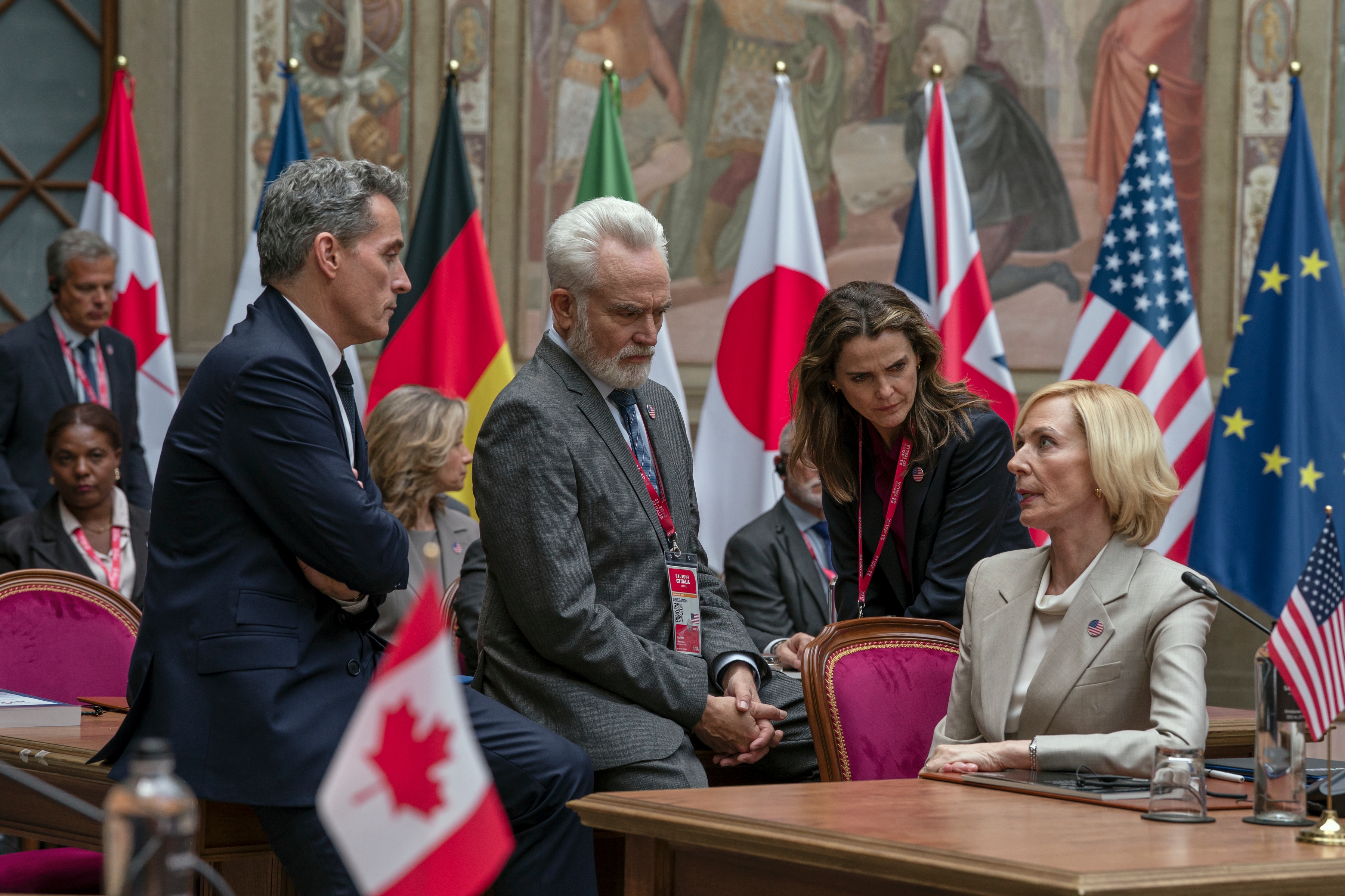 &Quot;Four People In Formal Attire Have A Serious Discussion At An International Political Meeting Room, Surrounded By Flags From Various Countries Including The Us, Canada, Japan, And The Eu. Conference Table With Microphones In Foreground.&Quot;